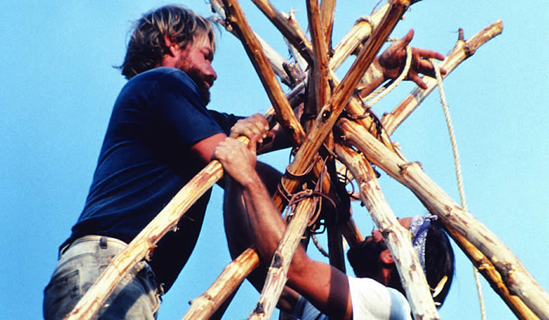 photograph looking up at two men tying wood poles together with blue sky behind them