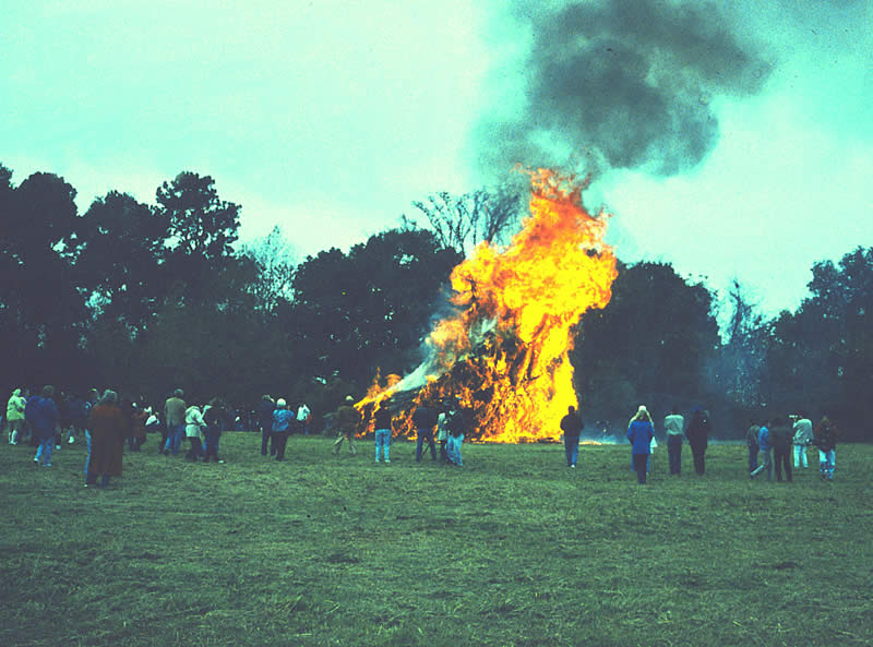 in the distance, a grass house engulfed in flames with billowing black smoke and people looking on