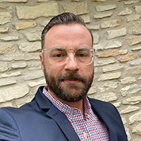 A photograph of a man outdoors wearing a blue suit and glasses