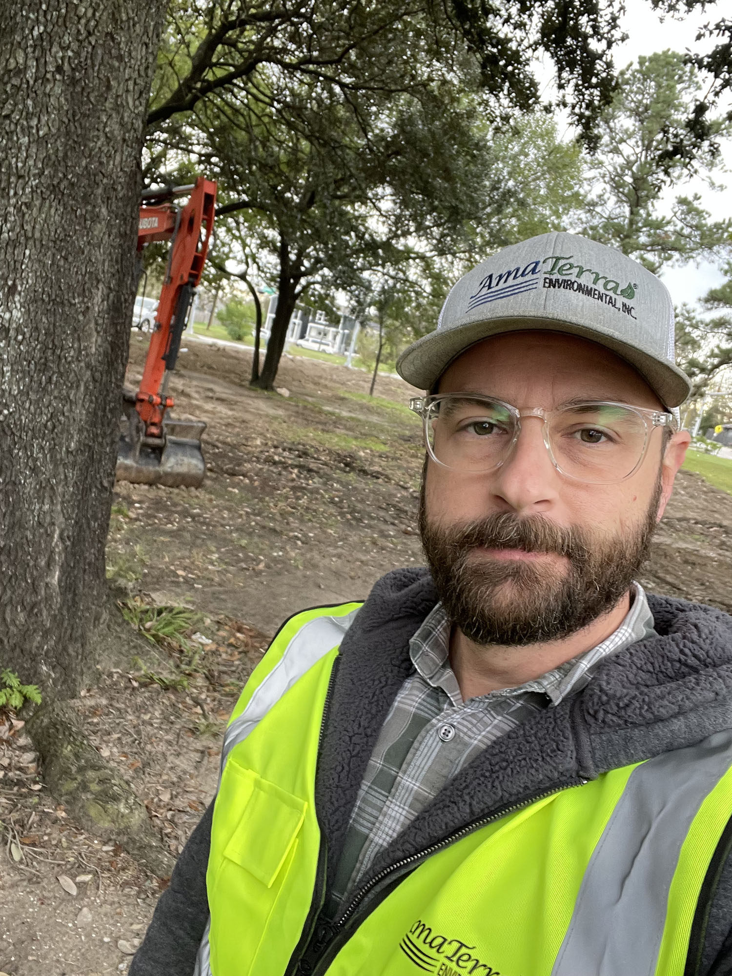 A photograph of a man wearing a ball cap and reflective vest with oak trees and bare dirt