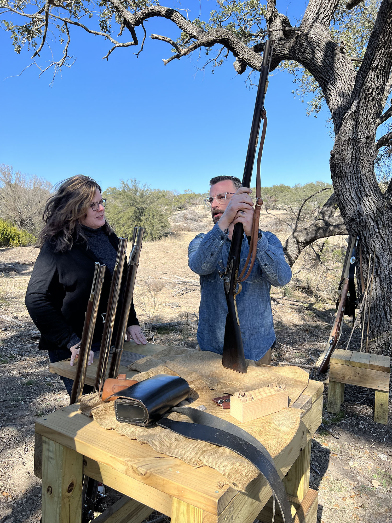 A photograph of a woman with long brown hair speaking with a man with short brown hair holding a musket with a live oak tree in the background