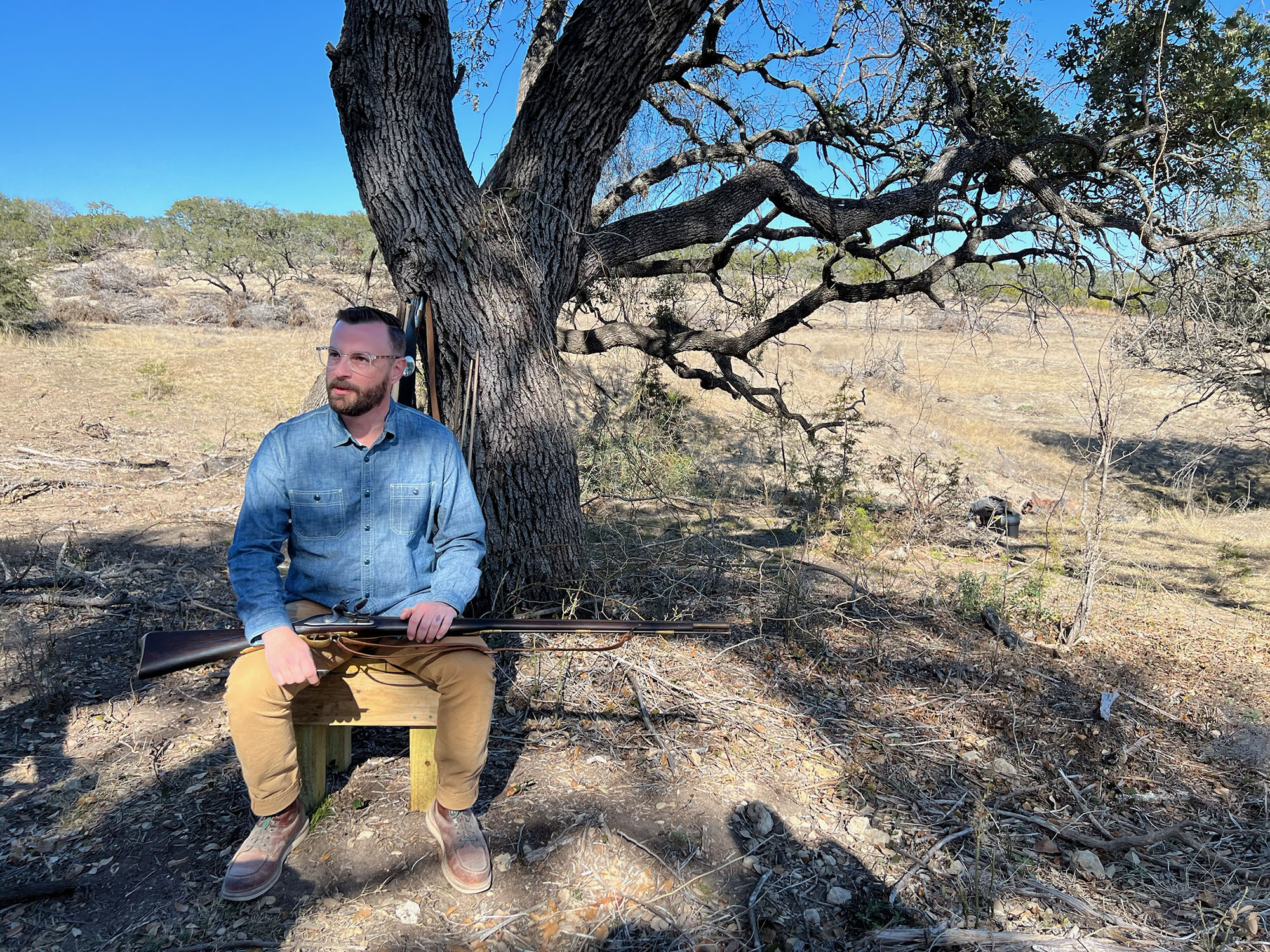 A photograph of a white man with brown hair sitting on a chair under a tree with a musket on his lap