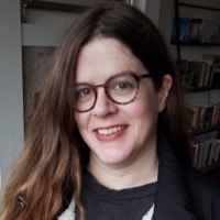 A photograph of a smiling woman wearing glasses with shelves behind her