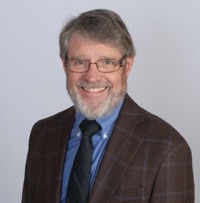 A formal photograph of a smiling man wearing glasses in front of a grey background