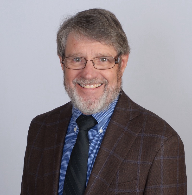 a white man in a suit with grey facial hair sitting in front of a grey wall