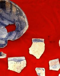 Photograph of broken grey colored pottery, some of it refit to form a portion of a bowl, on a red background.