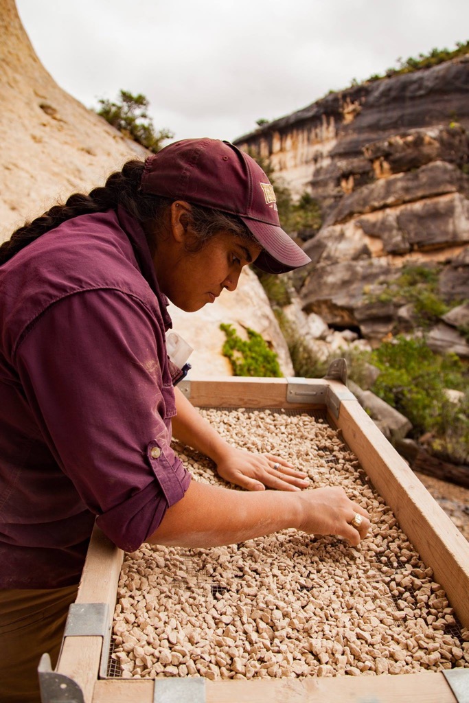 a latina looking through an archeological screen filled with small rocks in a desert canyon