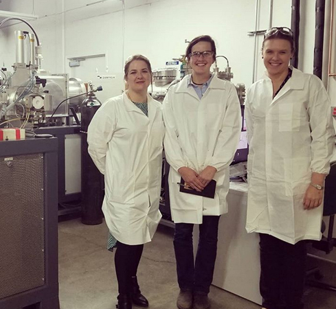 Photograph of three smiling women in white lab coats, with laboratory equipment in the background