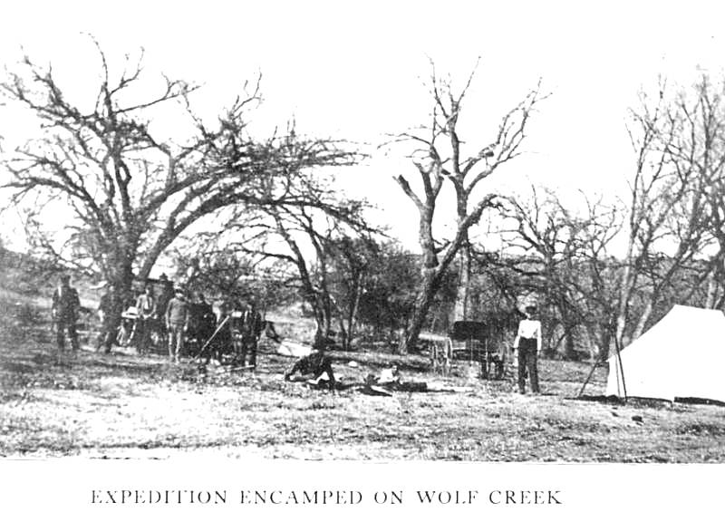 Old photo showing students, tent, and automobile in wooded area.