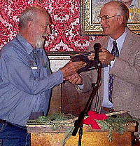 Photo of man in coat and tie handing a plaque to man dressed in blue denium.